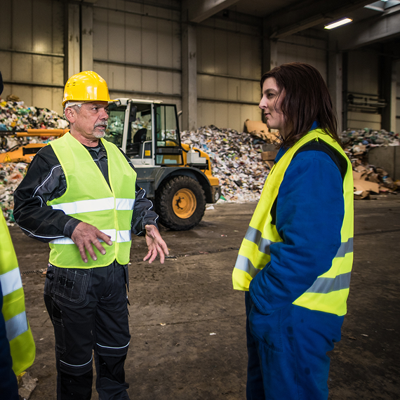 Two workers in high-visibility vests discuss in an industrial warehouse.